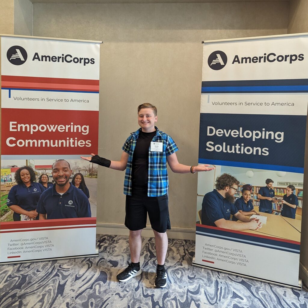 A young person poses in front of two AmeriCorps promotional signs at an event.